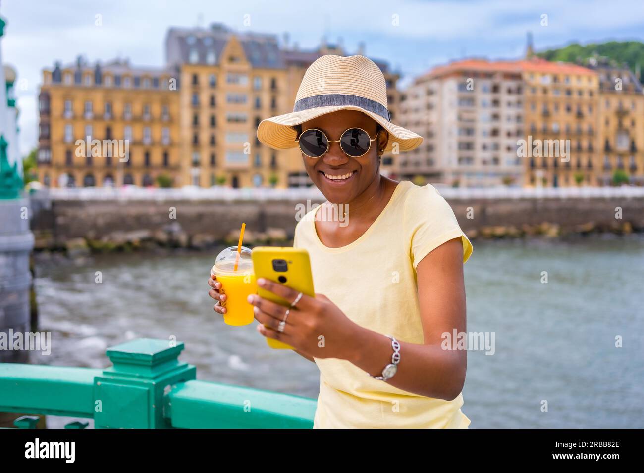 Schwarze ethnische Frau, die Orangensaft am Meer in der Stadt trinkt, Touristen, die den Sommer genießen Stockfoto