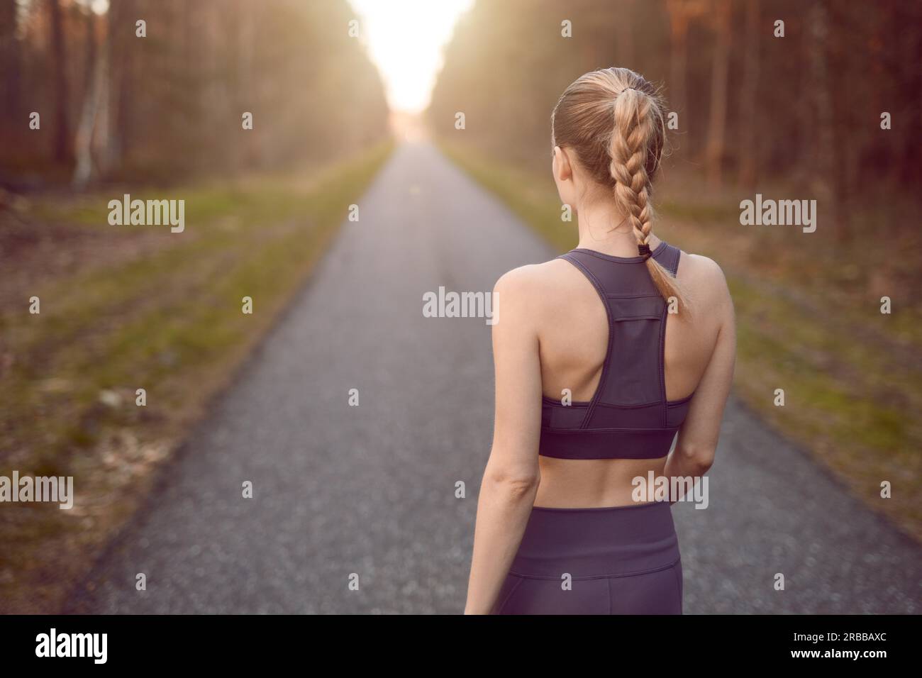 Sportlich gesunde junge Frau zu Fuß bei Sonnenaufgang entlang einer Landstraße durch einen dichten Wald in Richtung der Schein der Sonne am Ende zwischen den Bäumen in A Stockfoto