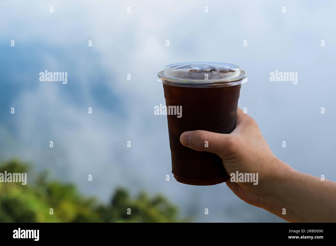 Halten Sie ein Glas Eiskaffee mit verschwommenem Berg- und Wolkenhintergrund in der Hand. Sommergetränk-Konzept. Stockfoto