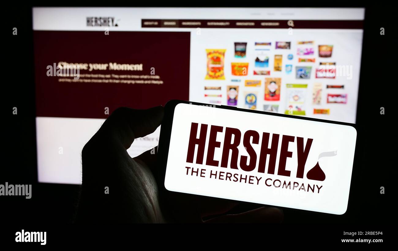 Person, die ein Mobiltelefon mit dem Logo des US-amerikanischen Lebensmittelunternehmens The Hershey Company auf dem Bildschirm vor der Unternehmenswebseite hält. Konzentrieren Sie sich auf das Display des Telefons. Stockfoto