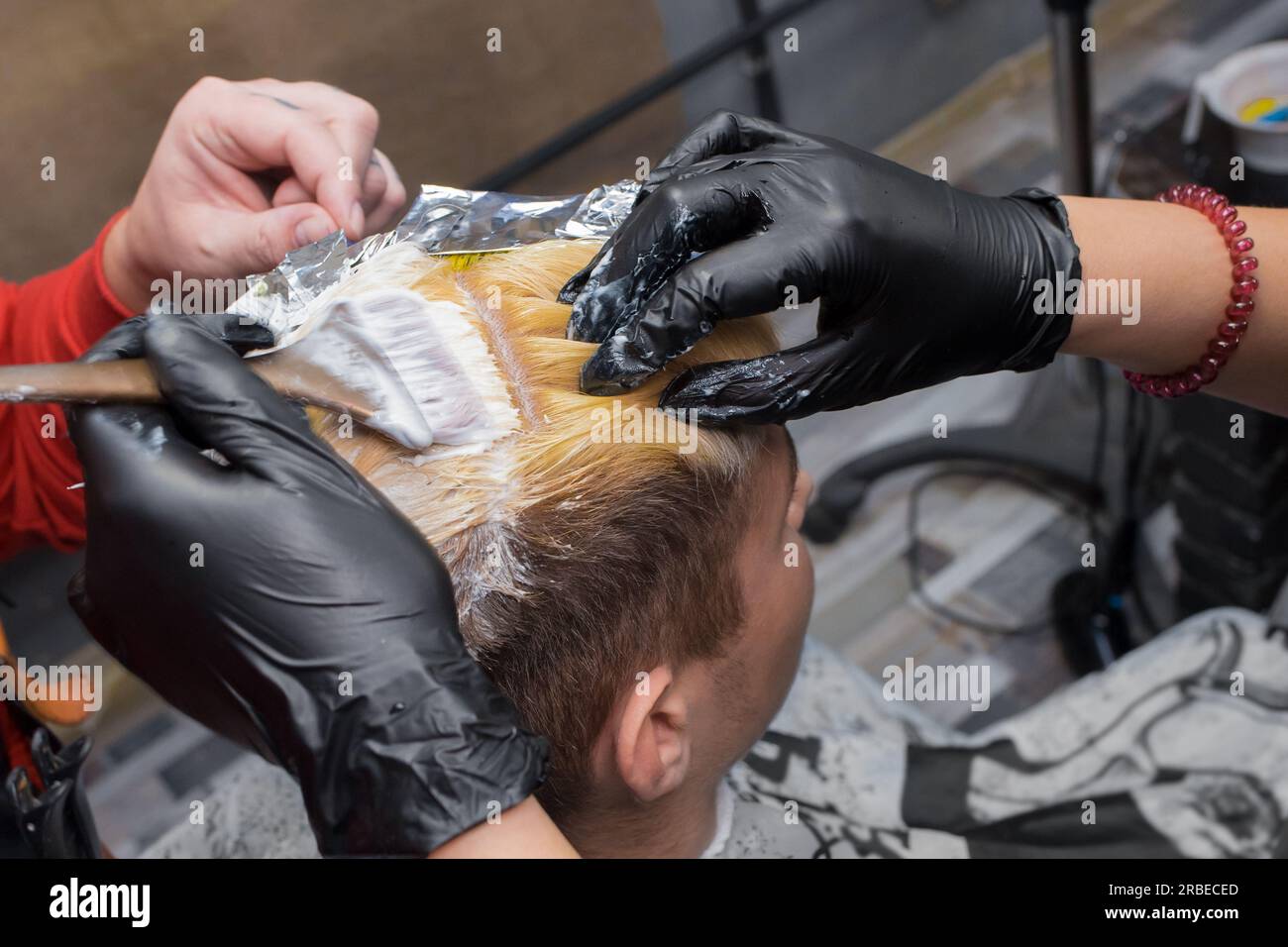 Hände einer Person in Handschuhen beim Färben von Männerhaaren mit einer Bürste bei der Arbeit in einem Friseursalon oder Friseur. Stockfoto