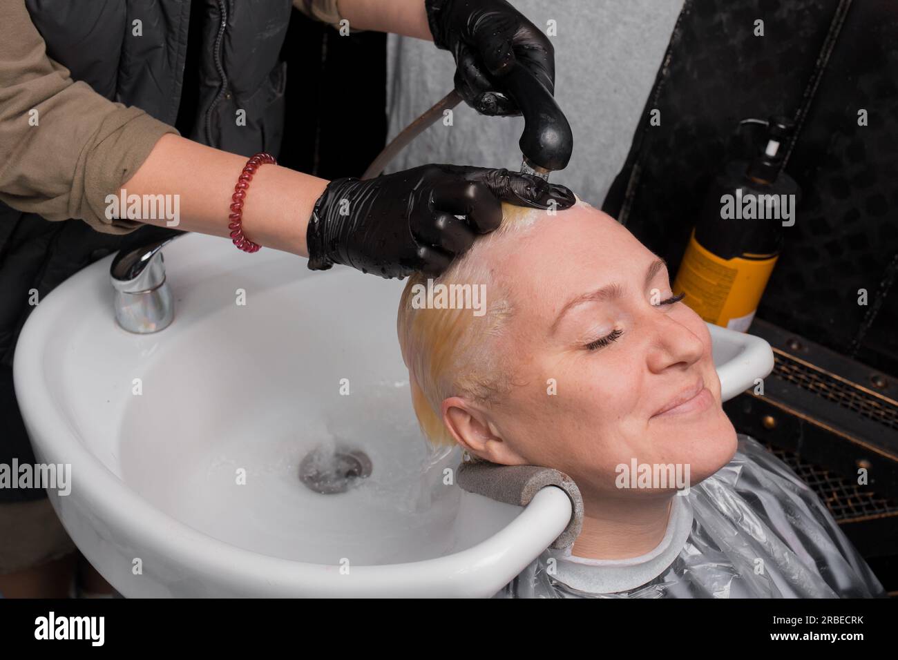 Eine lächelnde Erwachsene Weiße wäscht ihr Haar mit Wasser über dem Waschbecken, bevor sie in einem Friseur oder Friseur frisiert. Stockfoto