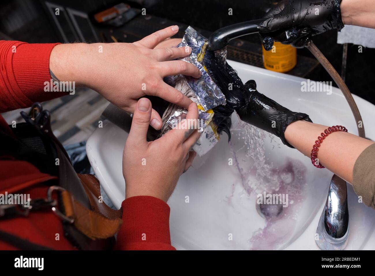 Die Hände eines Friseurs und eines Friseurs, die in einem Team arbeiten und sich mit Regen über dem Waschbecken waschen, nachdem sie einem Kunden, einem Mann im Salon, gefärbt haben. Stockfoto