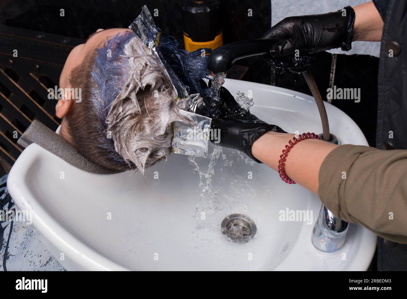 Die Hände eines professionellen Friseurs, ein Mädchen in Handschuhen, waschen sich die Farbe ab, während sie die Haare eines Klienten über einem Waschbecken mit Wasser färben, rai Stockfoto