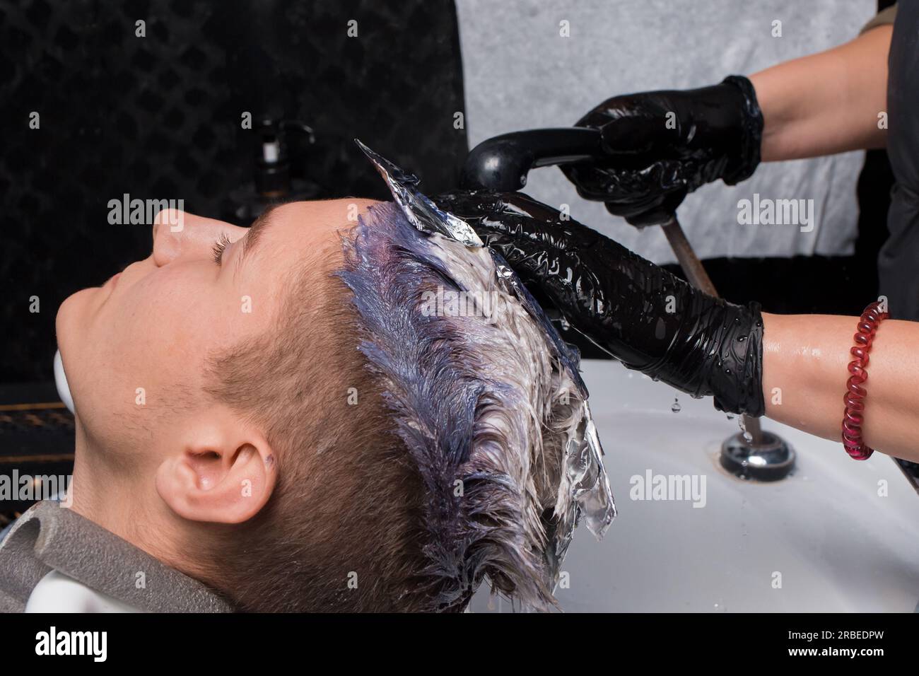 Die Hände eines Mädchens, eines Friseurarbeiters, das Handschuhe mit Regen trägt, Wasser wäscht die Farbe über dem Waschbecken ab, nachdem er die Haare eines Kunden gefärbt hat, eines Mannes bei Stockfoto