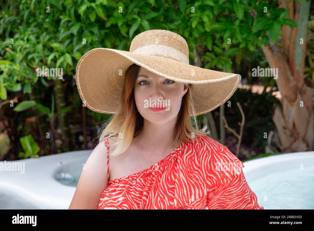 Wunderschöne weiße Frau, die ein auffälliges rotes Sommerkleid mit einem übergroßen Strohhut trägt, während sie direkt in die Kamera blickt Stockfoto