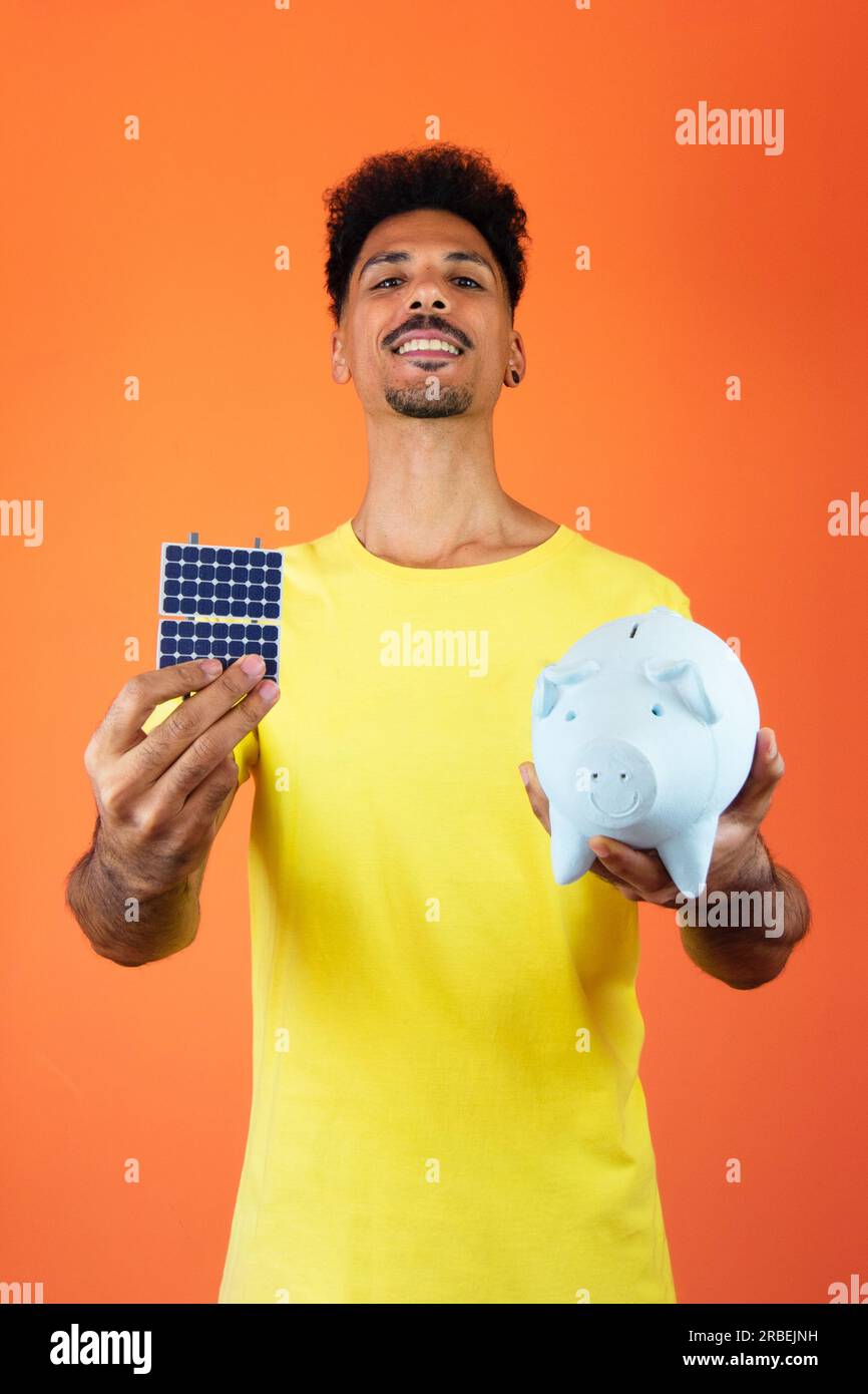 Black man hält eine Piggy Bank und Solarpaneel isoliert auf Orange. Stockfoto