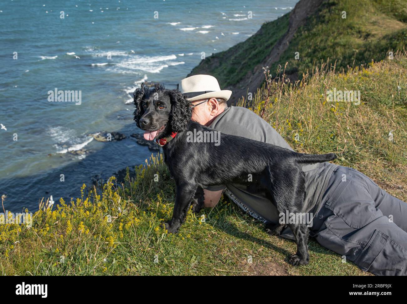 Ein älterer Mann mit einem Hut und einem schwarzen Hund, der über eine Klippe blickt Stockfoto