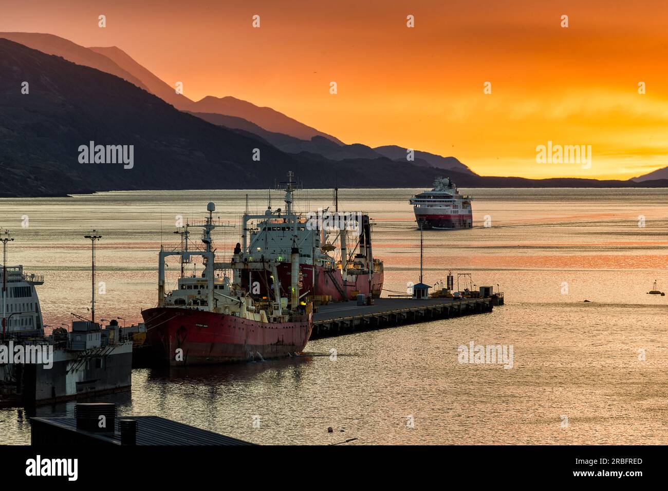 Sehr farbenfroher Sonnenaufgang im Hafen von Ushuaia, mit ein paar angedockten Booten im Vordergrund und ein weiteres nähert sich in der Ferne Stockfoto