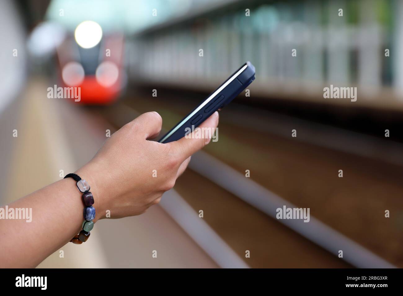Smartphone in der Hand einer Frau, die auf den ankommenden Zug am Bahnhof wartet. Pendlerzugpassagier, Online-Zahlung für die Reise im Sommer Stockfoto