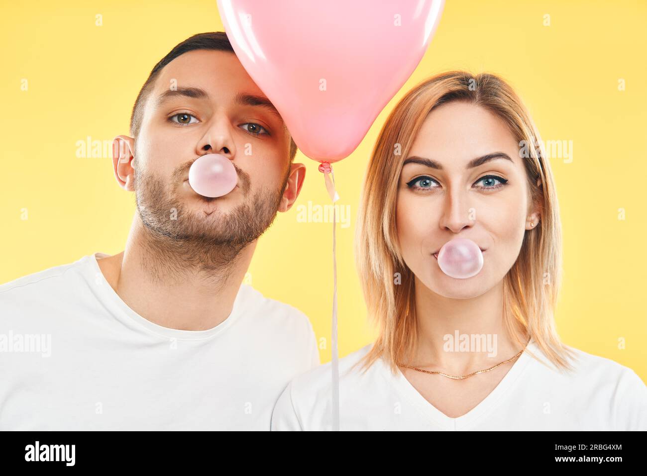 Junges Paar bläst Blase mit Kaugummi und hält rosa Luftballon auf gelbem Hintergrund Stockfoto