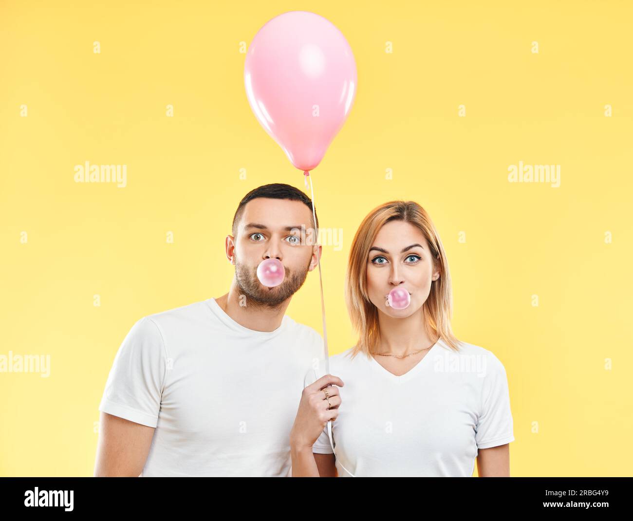 Junges Paar bläst Blase mit Kaugummi und hält rosa Luftballon auf gelbem Hintergrund Stockfoto