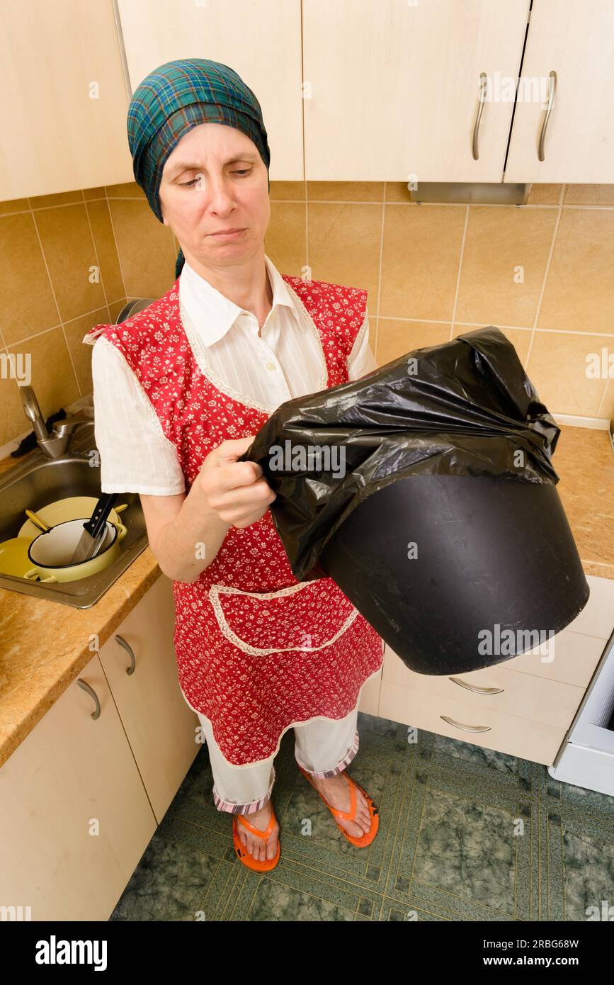 Eine Frau mit einem Schal auf dem Kopf und einer roten Schürze schaut in einen schwarzen Mülleimer mit einem Müllsack in der Küche. Sie ist sehr beunruhigt Stockfoto