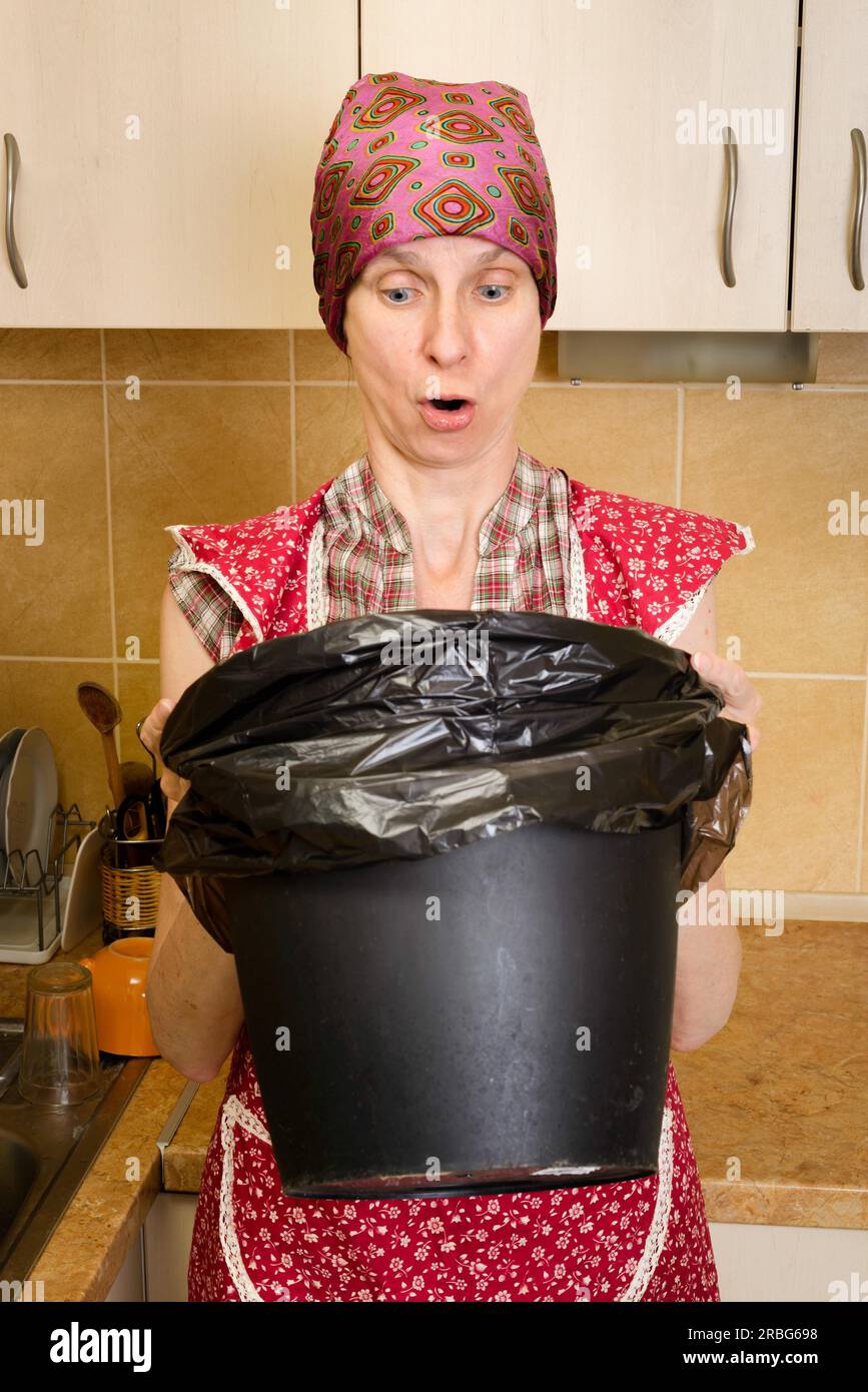 Eine Frau mit einem Schal auf dem Kopf und einer roten Schürze schaut in einen schwarzen Mülleimer mit einem Müllsack in der Küche. Sie ist sehr beunruhigt Stockfoto