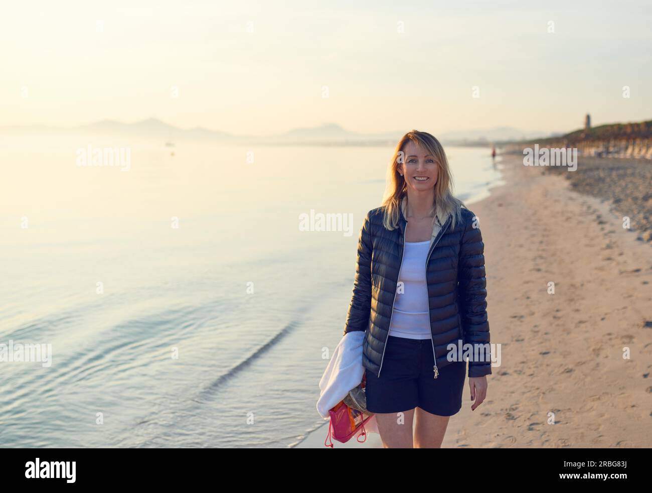 Fröhliche blonde Frau, die bei Sonnenuntergang am Strand entlang spaziert Stockfoto