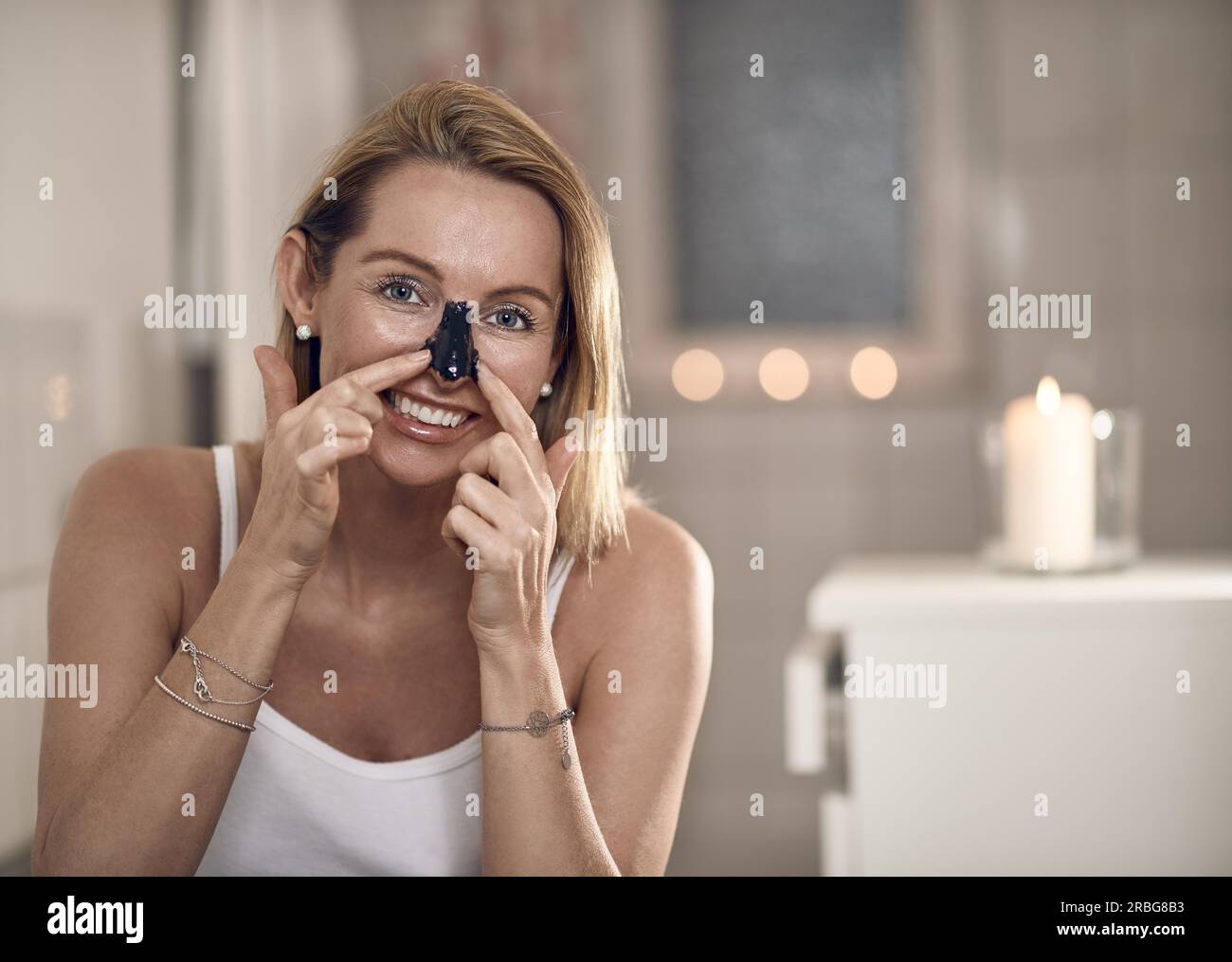 Attraktive blonde Frau mittleren Alters, die eine Anti-Aging-Gesichtsmaske auf ihre Nase in einem Badezimmer mit brennenden Kerzen in einem Konzept von Schönheit, Hautpflege Stockfoto