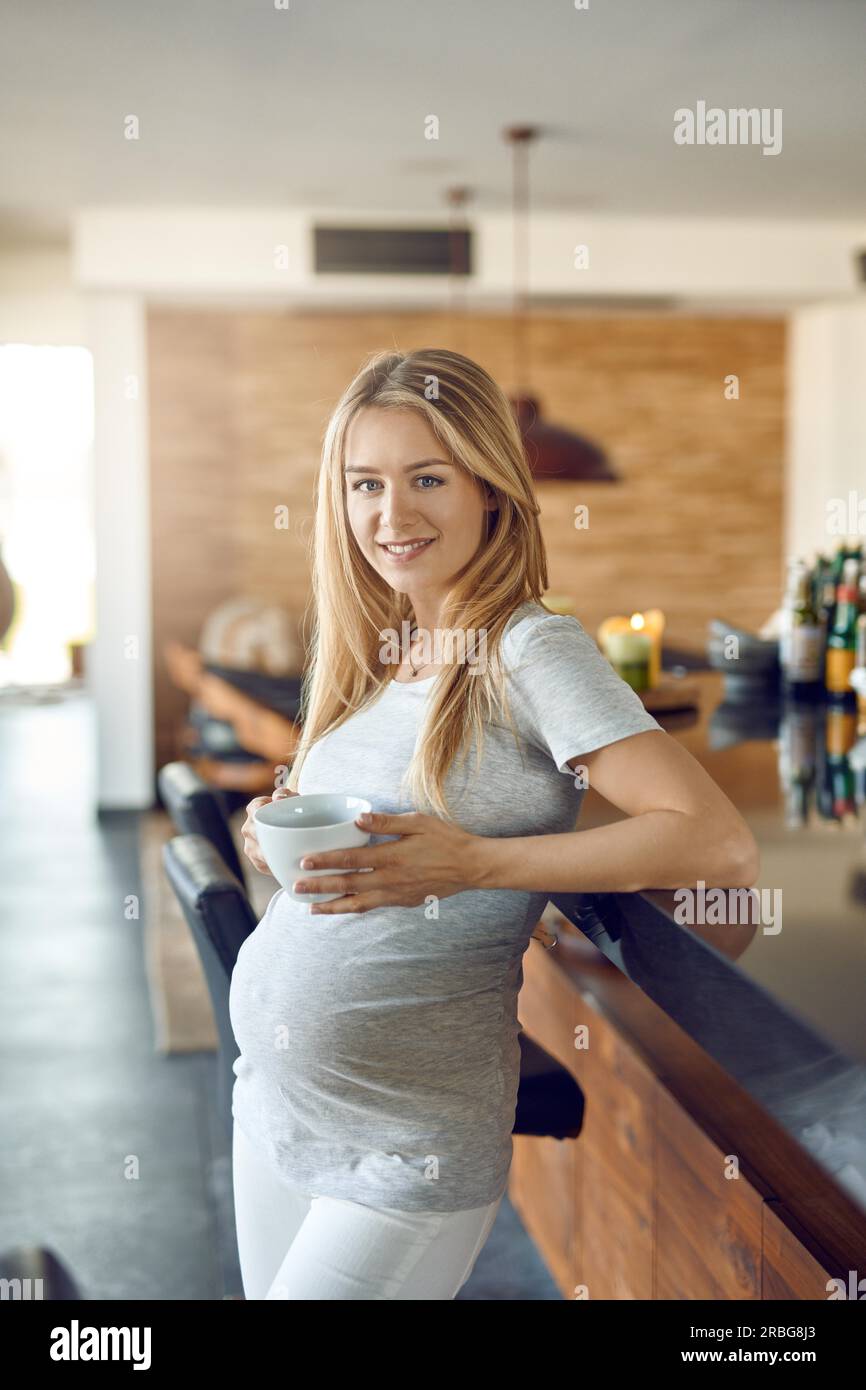 Lächelnde, fröhliche, schwangere Frau, die sich entspannt, sich mit einer Tasse heißem Tee an einer Bar lehnt und ihr Baby in der Hand hält, während sie sich mit ihr verbindet Stockfoto