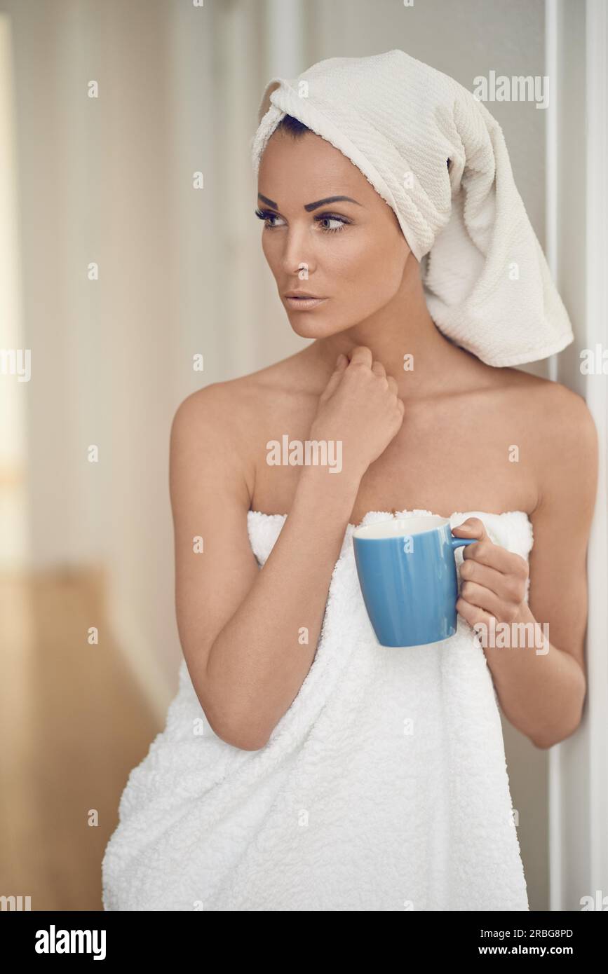 Eine hübsche Frau mittleren Alters, eingewickelt mit frischen, sauberen, weißen Handtüchern um ihren Kopf und Körper, die sich an eine Innenwand lehnt, mit einer Tasse Getränk in einem Stockfoto