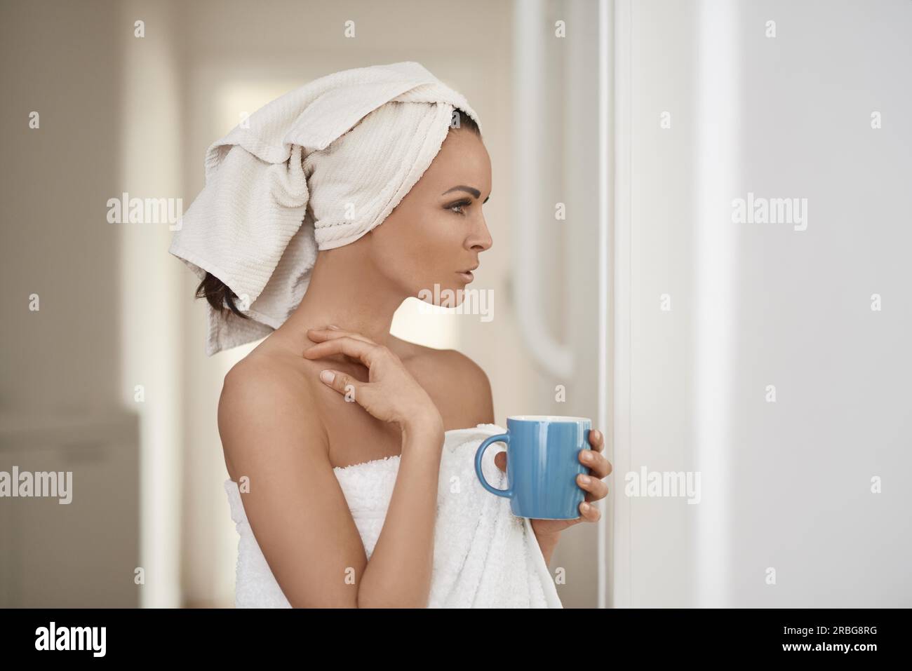 Eine hübsche Frau mittleren Alters, eingewickelt mit frischen, sauberen, weißen Handtüchern um ihren Kopf und Körper, die sich an eine Innenwand lehnt, mit einer Tasse Getränk in einem Stockfoto
