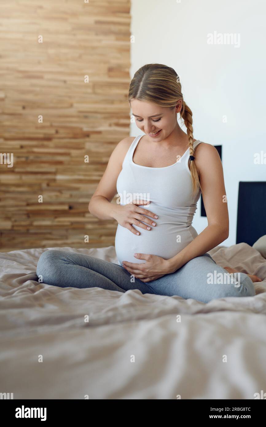 Glückliche, schwangere Frau, die sich mit ihrem ungeborenen Kind verbindet, sitzt auf einem Bett und wiegt ihr Baby mit den Händen nach unten, mit einem zärtlichen Lächeln von Stockfoto