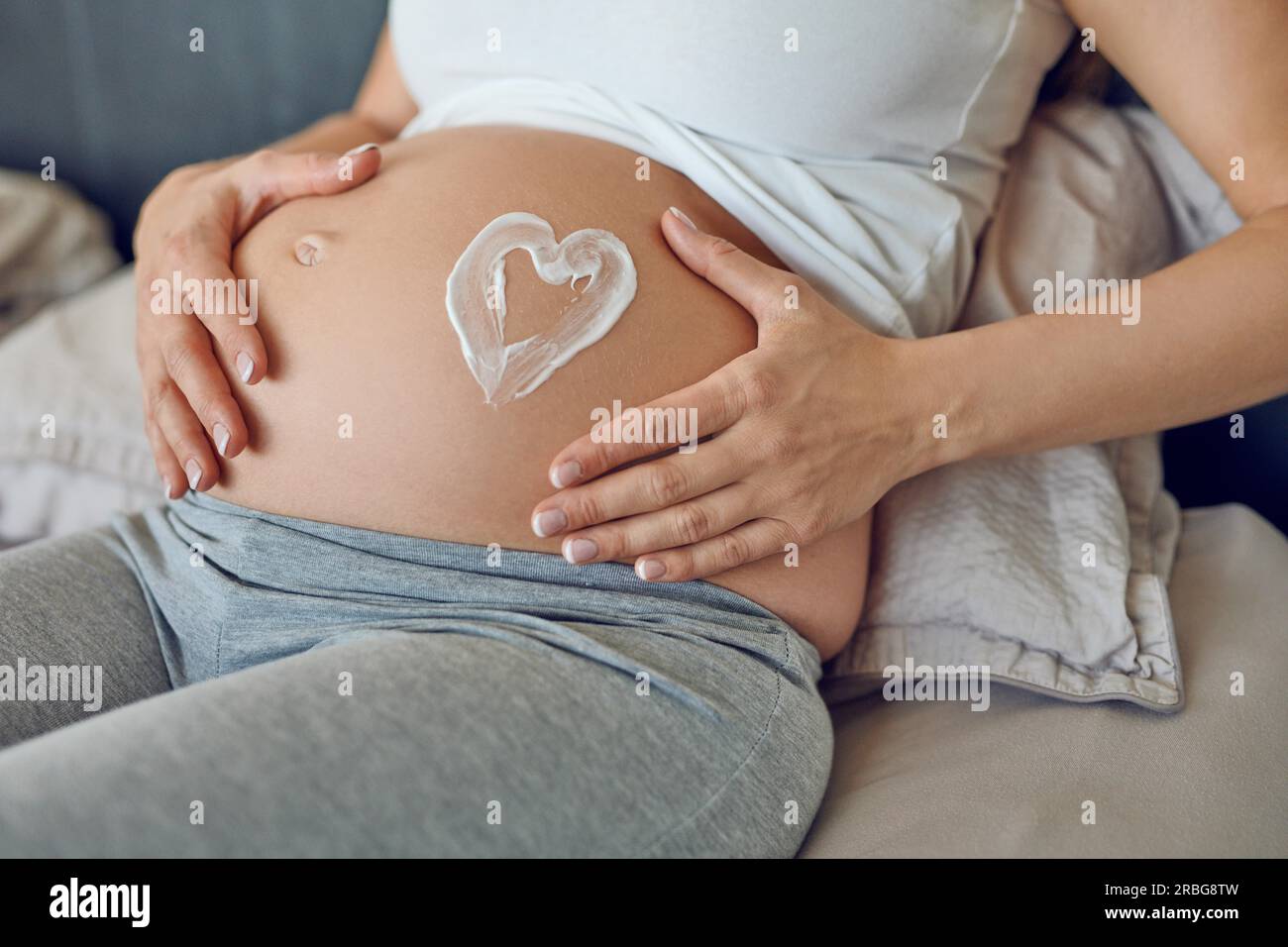 Eine junge schwangere Frau malt ein Herz in feuchtigkeitsspendender Creme auf ihre Babybrücke, während sie ihren Bauch in den Händen hält, während sie sich in der Nähe eines Bettes entspannt Stockfoto
