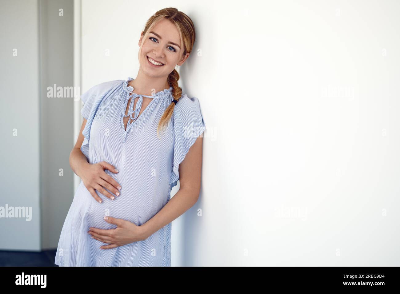 Glückliche schwangere junge Frau, die ihren Bauch mit ihren Händen umklammert, während sie sich mit ihrem ungeborenen Kind verbindet und an einer weißen Wand posiert und dabei lächelt Stockfoto