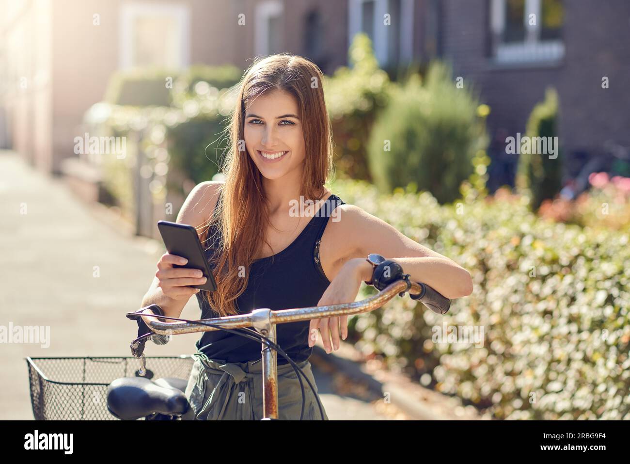 Wunderschöne junge Frau mit langen Haaren in einem halblangen Porträt draußen in der Stadt, die sich mit einem modernen Smartphone auf ein altes Fahrrad lehnt Stockfoto Wunderschöne junge Frau mit langen Haaren in einem halblangen Porträt draußen in der Stadt, die sich mit einem modernen Smartphone auf ein altes Fahrrad lehnt Stockfoto