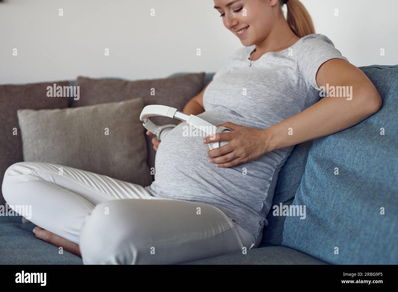 Lächelnd glücklich schwangere junge Frau, die Musik mit Kopfhörern auf Ihr ungeborenes Baby, indem Sie einen Stereokopfhörer an ihrem Bauch wie Sie sitzt Stockfoto