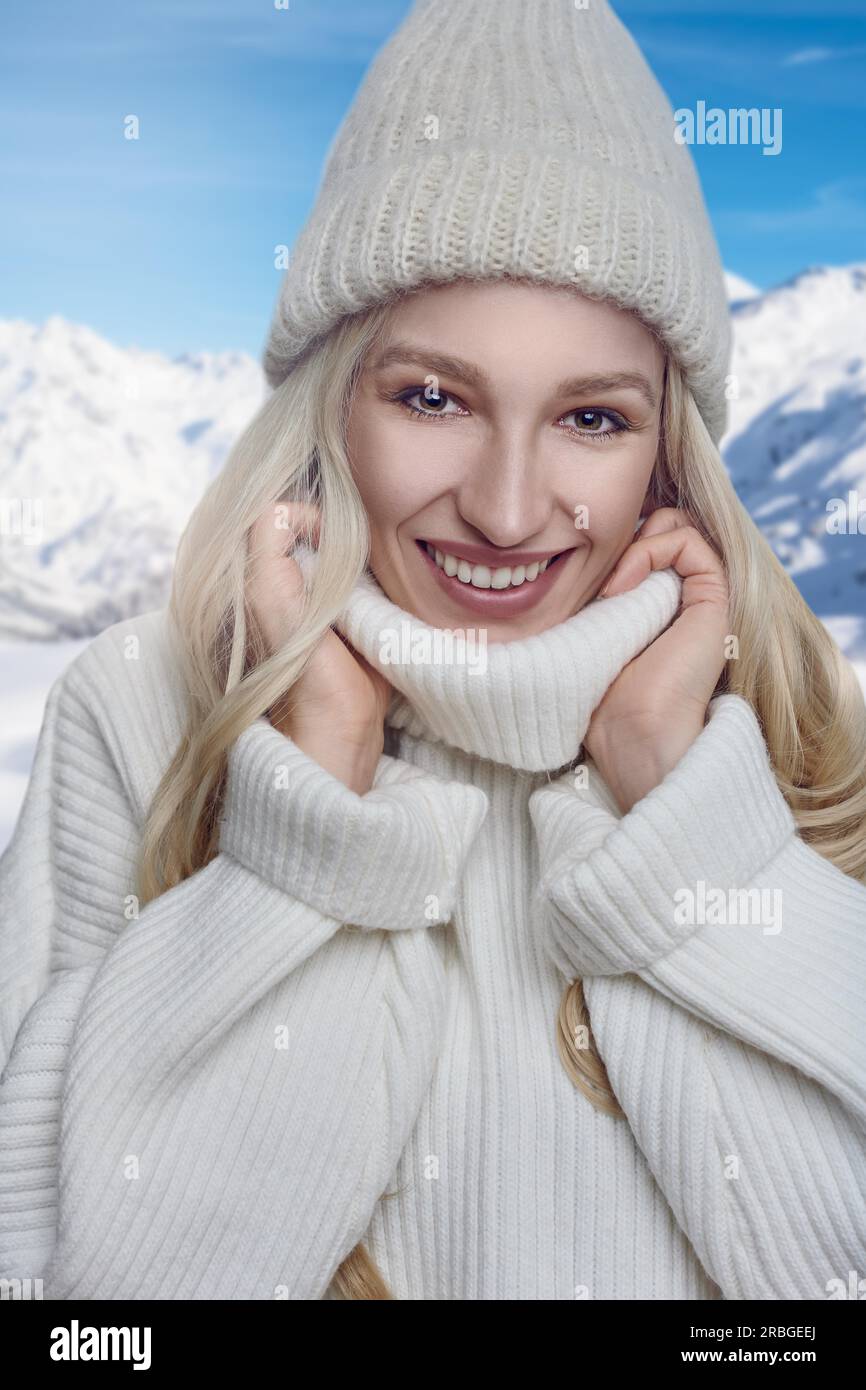 Eine attraktive blonde Frau in einem warmen Wollpullover mit Polokragen und Strickmütze, die sich im Freien mit einem fröhlichen Freund in den Kragen kuschelt Stockfoto