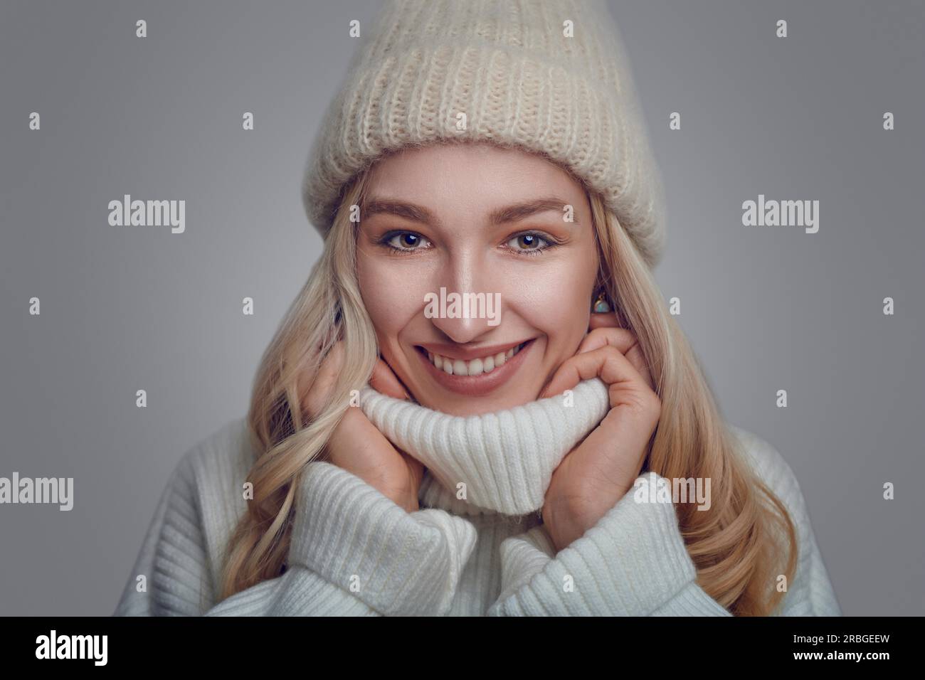 Attraktive blonde Frau in einem warmen Wollpullover mit Polokragen und Strickmütze, die vor einem grauen Hintergrund steht und sich mit einem in den Kragen kuschelt Stockfoto