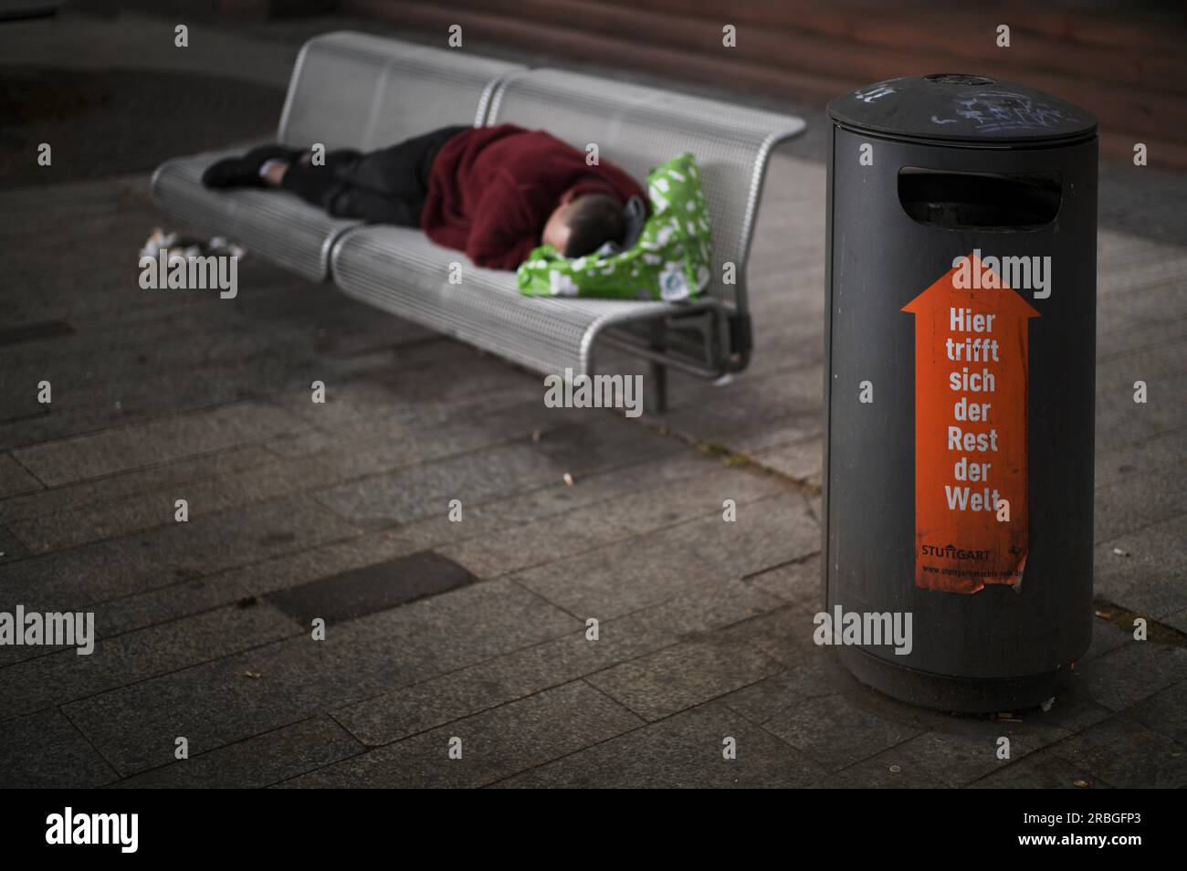 Obdachloser, Berber, der auf der Parkbank liegt, schläft, vor ihm Müllcontainer mit Schriftzug TRIFFT HIER AUF DEN REST DER WELT, Stuttgart Stockfoto