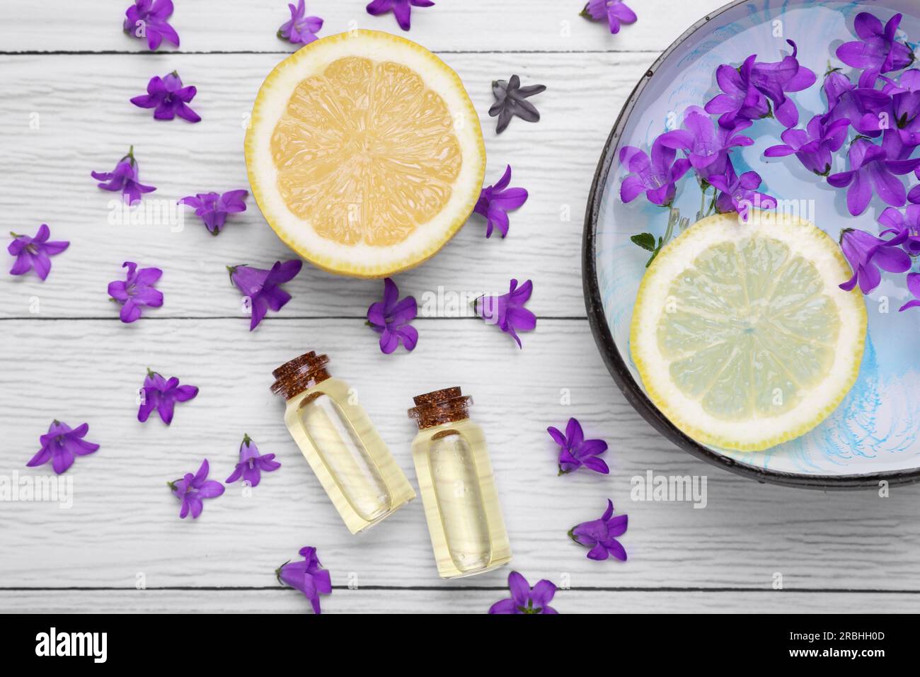 Schüssel mit Wasser, Zitrone, violetten Blumen und Flaschen mit ätherischen Ölen auf weißem Holztisch, flach liegend. Spa-Zusammensetzung Stockfoto