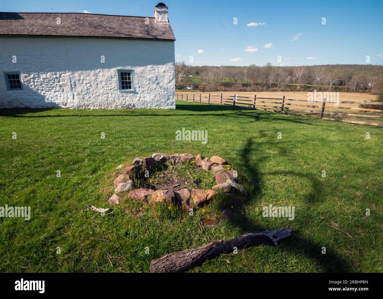 Koloniales amerikanisches Dorf mit idyllischem Häuschen aus weißem Stein in grünem Gras und einer Feuerstelle aus einem Steinring im Vordergrund. Weide und blauer Himmel Stockfoto