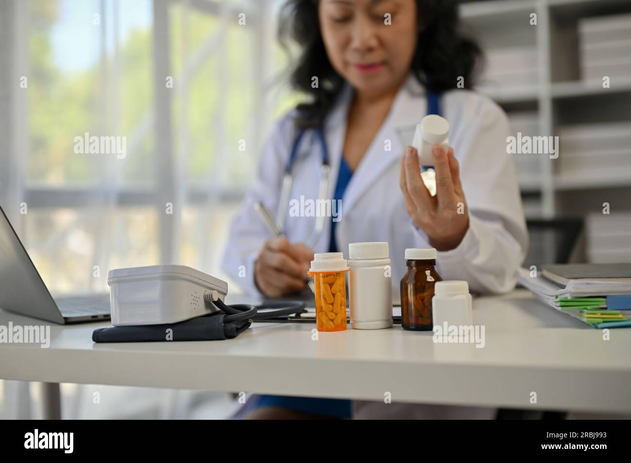 Nahaufnahme von Arzneimittelflaschen befindet sich auf einem Arztpraxistisch. Eine Ärztin, die an ihrem Schreibtisch in einem Untersuchungsraum ein Rezept ausstellt. Stockfoto