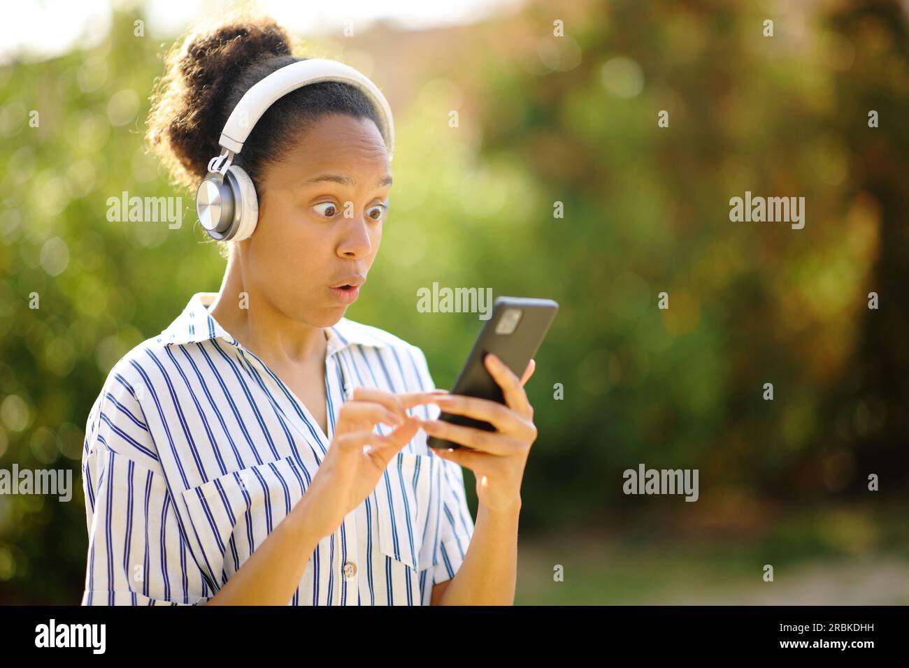 Eine überraschte schwarze Frau mit Kopfhörern, die in einem Park Musik am Telefon hört Stockfoto