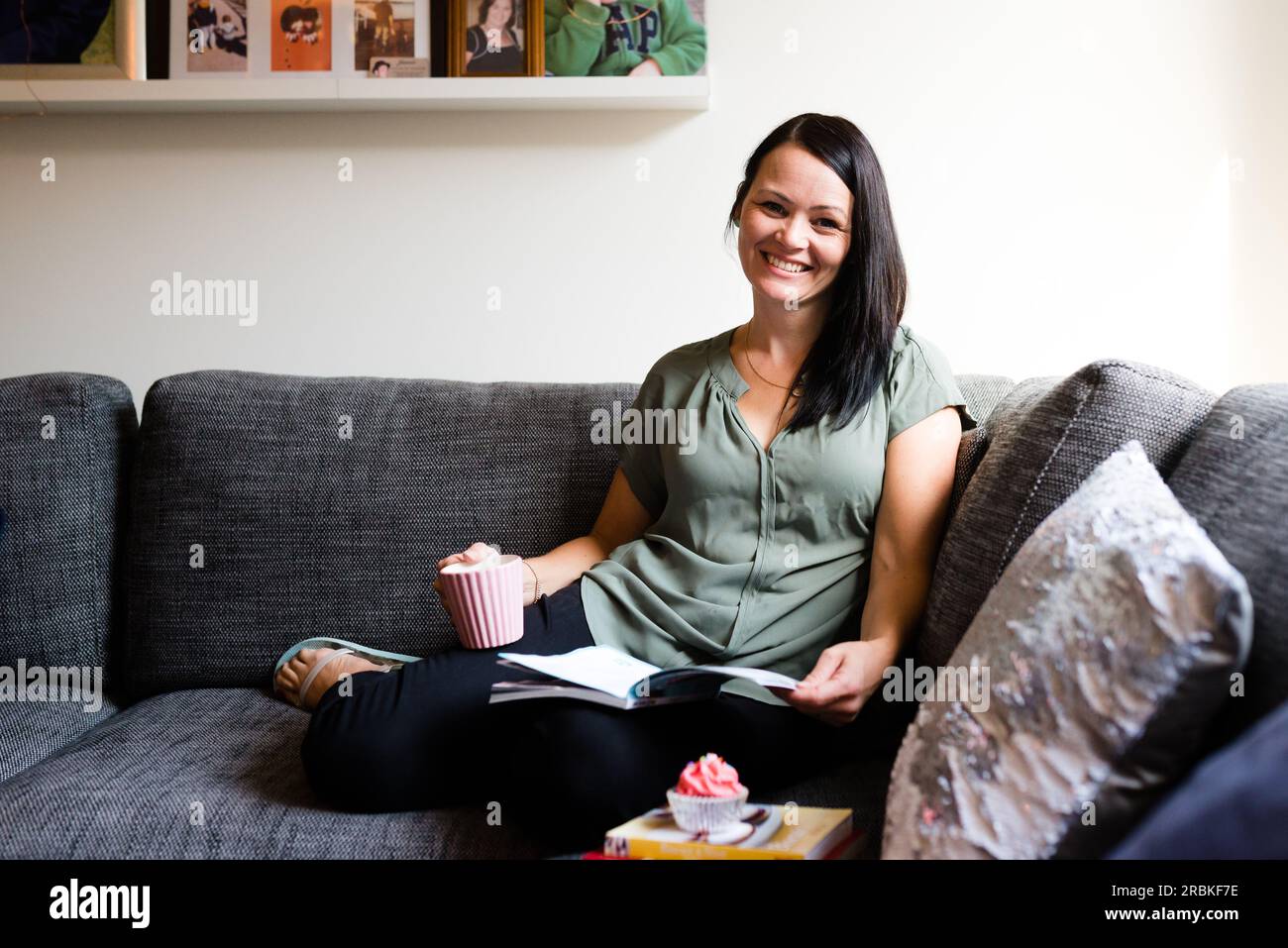 Eine Frau lächelt und liest mit einem Cupcake auf dem Sofa Stockfoto