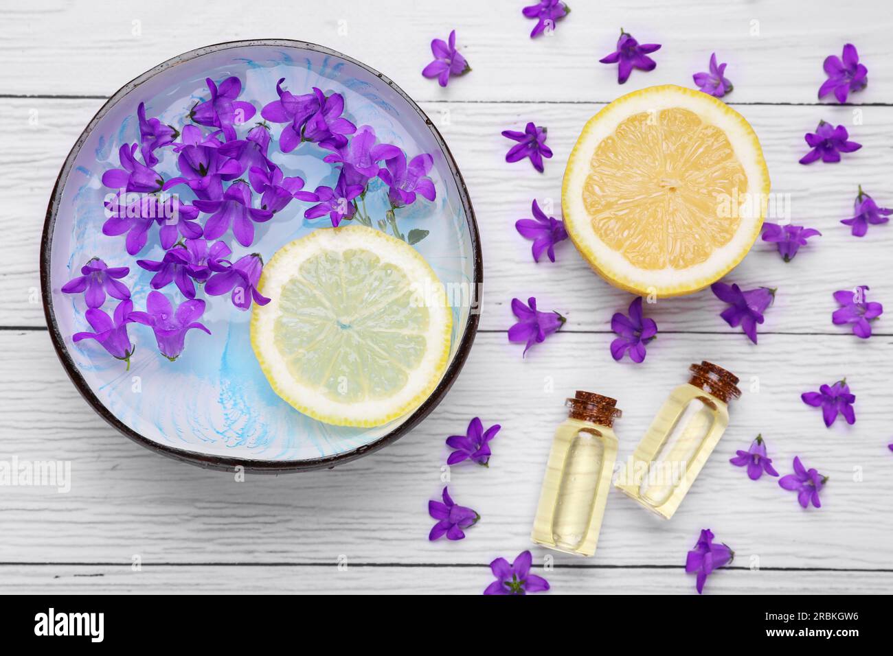 Schüssel mit Wasser, Zitrone, violetten Blumen und Flaschen mit ätherischen Ölen auf weißem Holztisch, flach liegend. Spa-Zusammensetzung Stockfoto