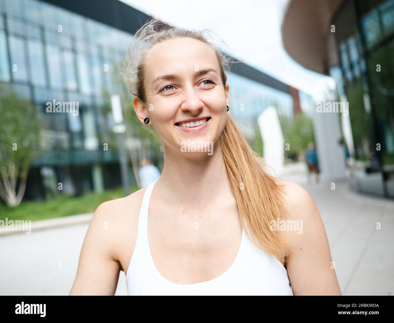 Junge blonde Frau lächelt draußen am sonnigen Sommertag mit urbanem Hintergrund Stockfoto
