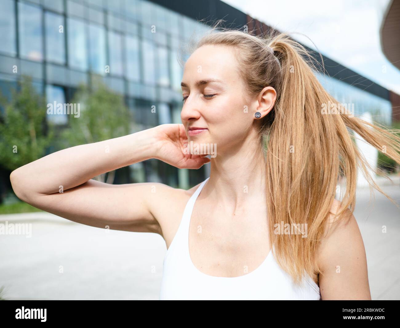 Eine junge blonde Frau glättet ihr Haar im Freien an sonnigen Sommertagen mit urbanem Hintergrund Stockfoto