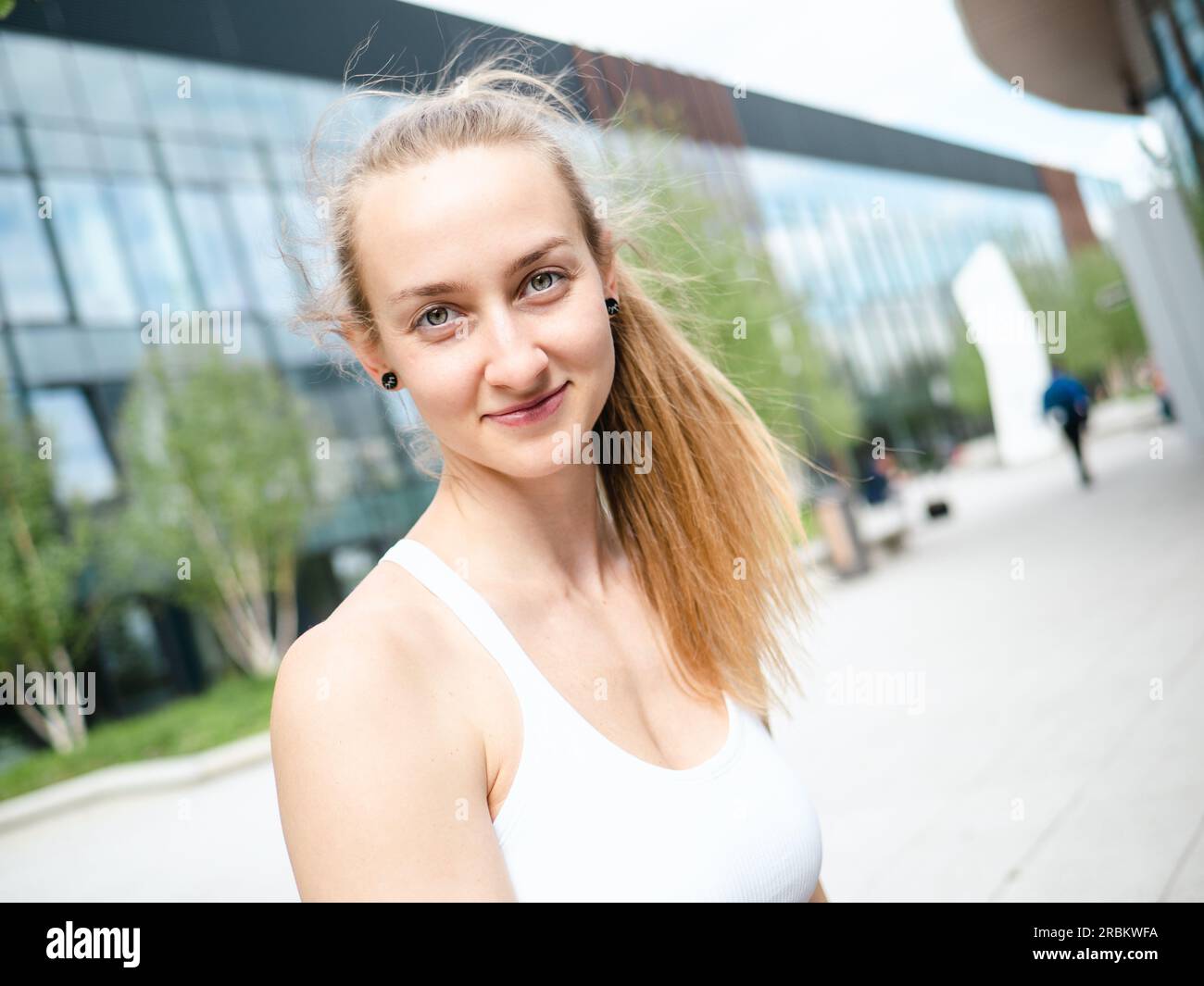 Junge blonde Frau draußen am sonnigen Sommertag mit urbanem Hintergrund, die in die Kamera schaut Stockfoto