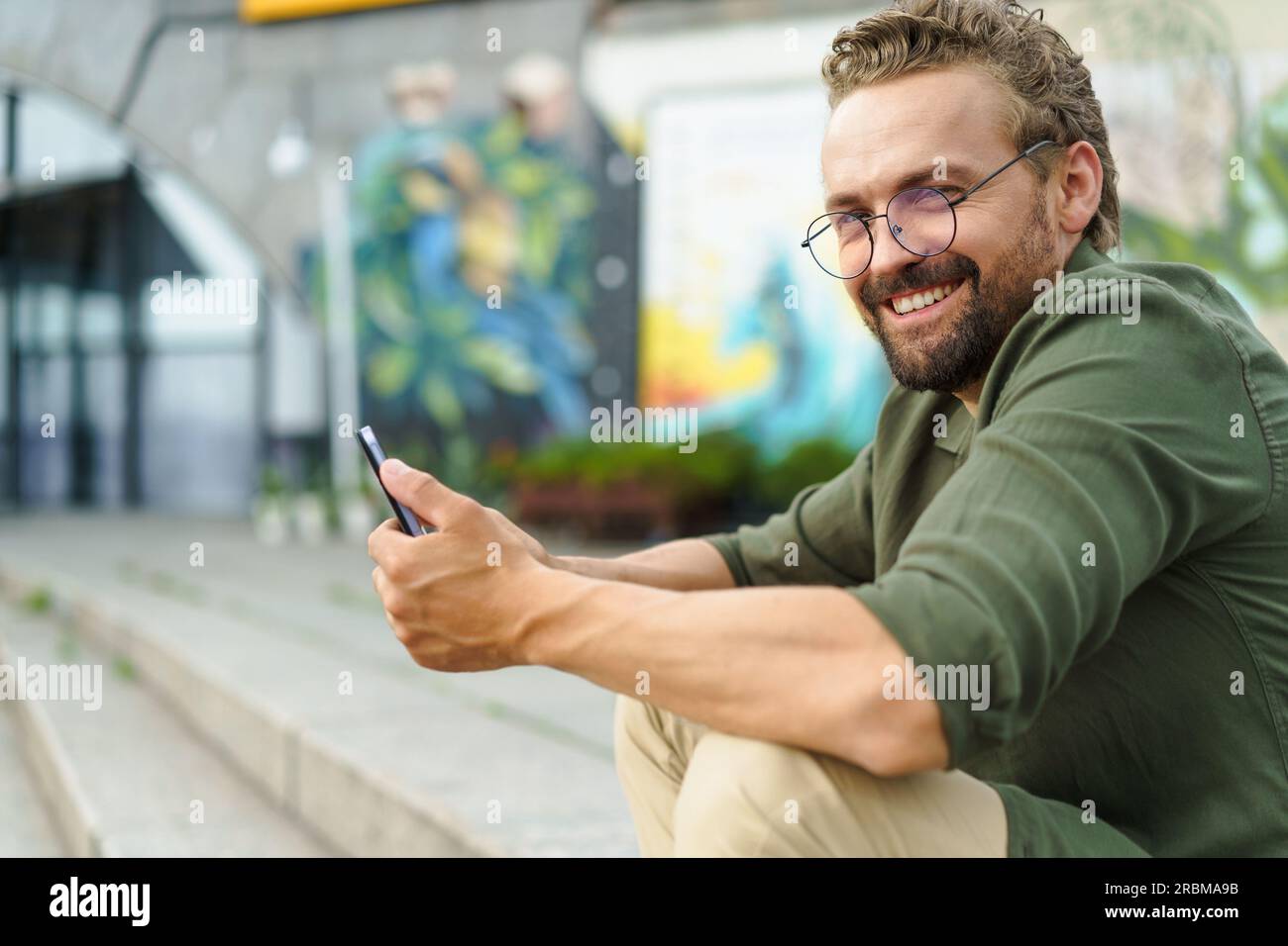 Ein Mann sitzt auf einer Steintreppe auf der Straße und benutzt einen Pocket PC. Mit seinem kompakten und tragbaren Gerät in der Hand kombiniert er mühelos Technologie und Umgebung. Der urbane Hintergrund verleiht der Szene einen modernen Touch. Hochwertiges Foto Stockfoto
