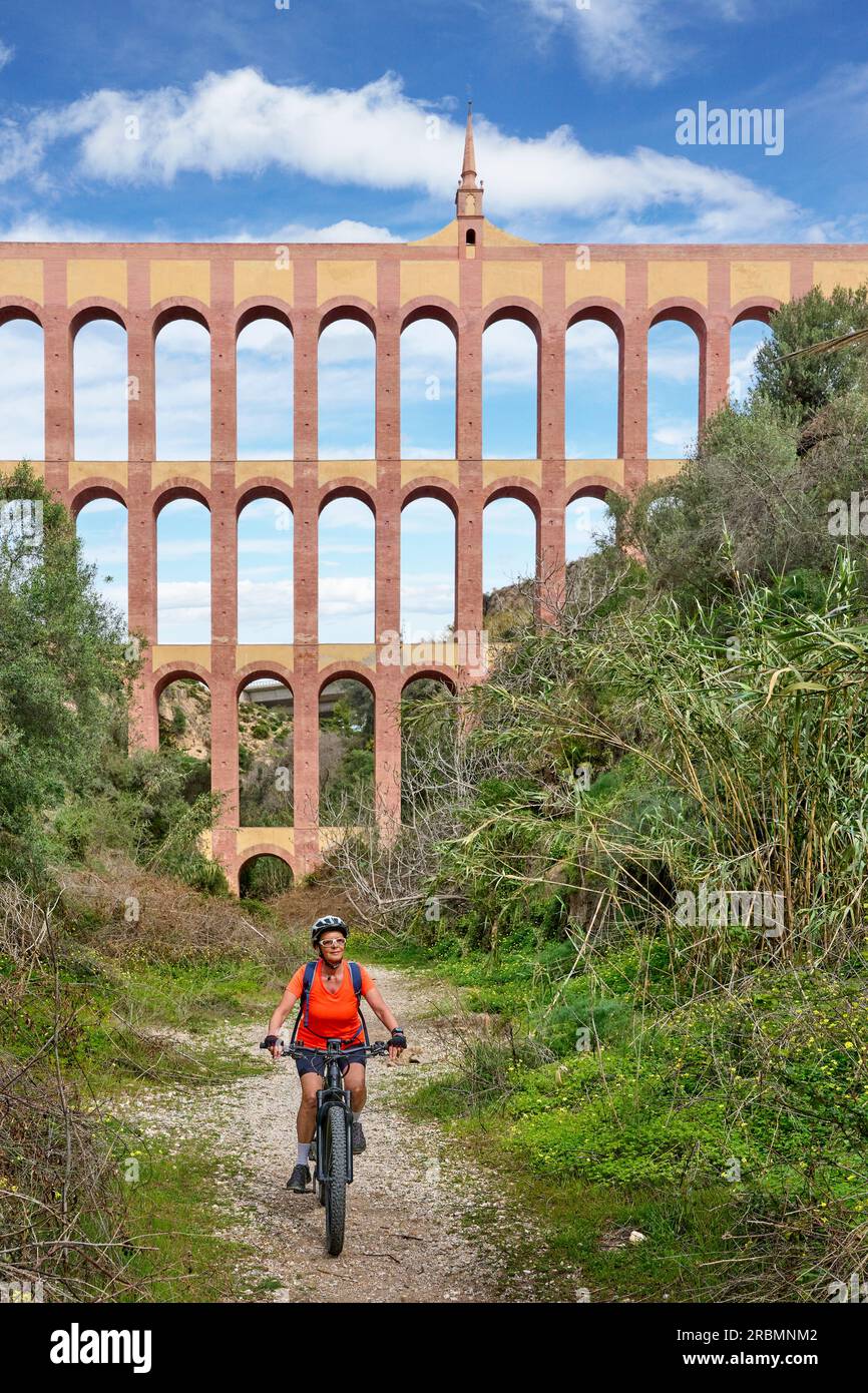 Nette Frau, die mit ihrem E-Mountainbike unter dem Aquädukt von Aguila in der Sierra de Tejada bei Nerja, Andalusien, Spanien, radelt Stockfoto