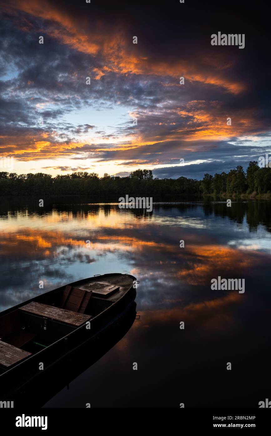 Ein See bei Sonnenuntergang, ein Boot davor. Orangefarbene Wolken am Himmel, die sich im See spiegeln. Rhein-Neckar-Region, Deutschland. Stockfoto