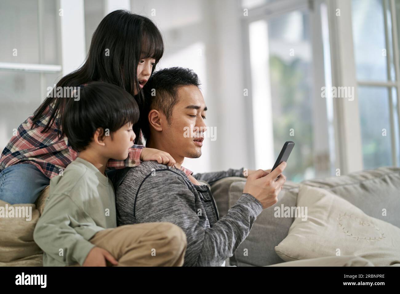 Junger asiatischer Vater und zwei Kinder, die zu Hause auf dem Familientisch sitzen und zusammen auf ein Handy schauen Stockfoto