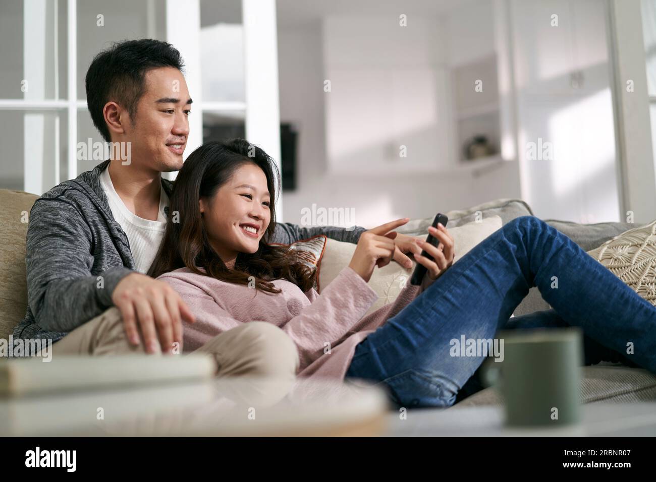 Glückliches junges asiatisches Paar, das zu Hause auf der Familientischcouch sitzt und sich Handyfotos ansieht Stockfoto