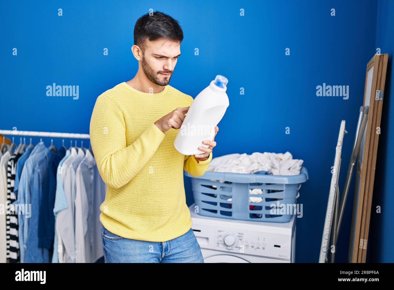 Junger hispanischer Mann liest Waschmitteletikett mit entspanntem Ausdruck in der Waschküche Stockfoto