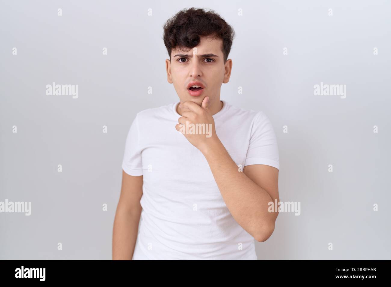 Junger nicht binärer Mann, der ein legeres weißes T-Shirt trägt, das fasziniert von Unglauben, Überraschung und erstaunlichem Ausdruck mit Händen am Kinn aussieht Stockfoto