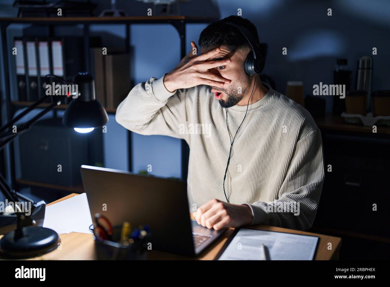 Ein junger, gutaussehender Mann, der nachts mit einem Computer-Laptop arbeitet und in einen Schock blickt, Gesicht und Augen mit der Hand verdeckt, durch Finger blickt und sich schämt Stockfoto