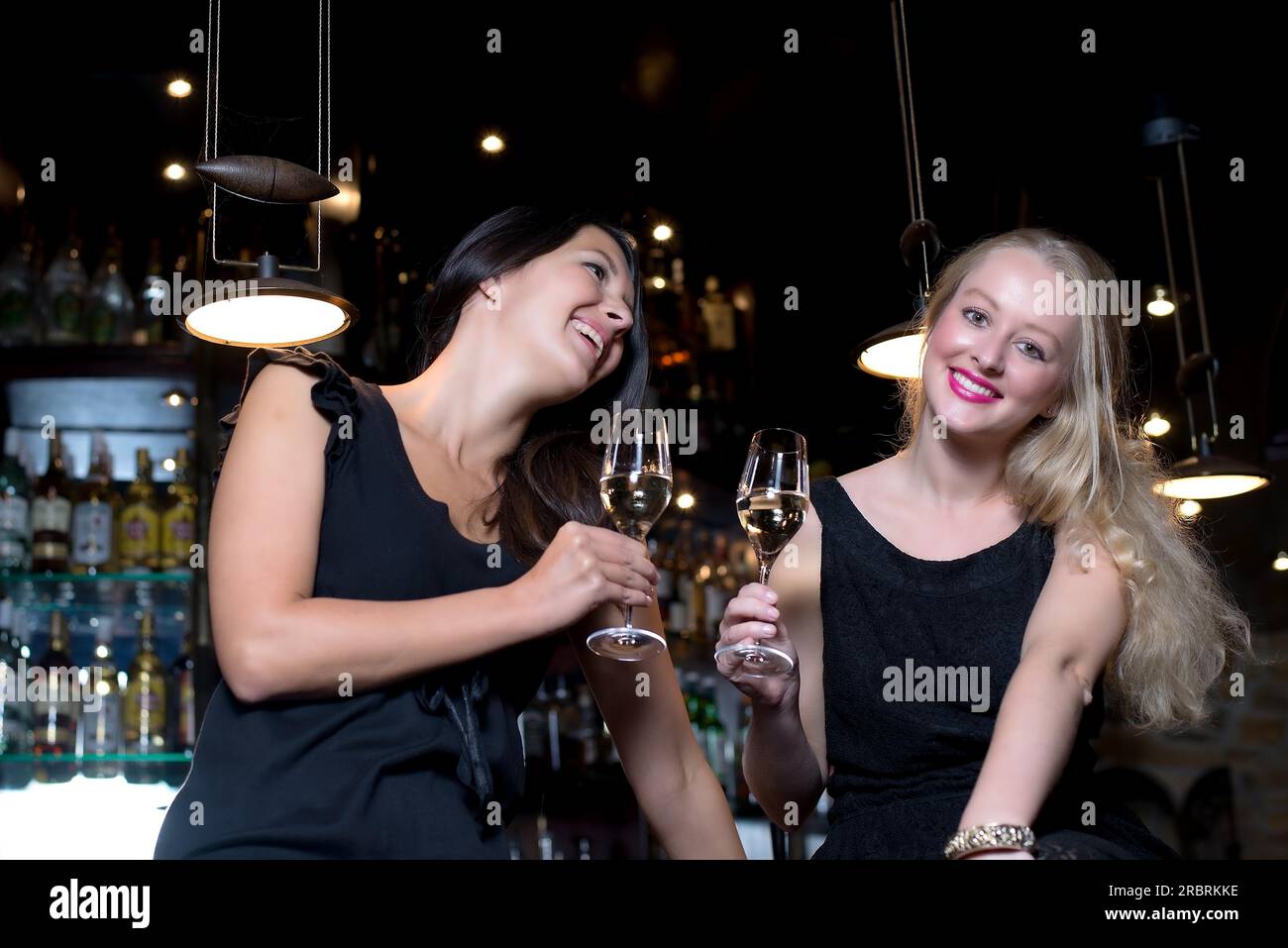 Zwei wunderschöne junge Frauen, die elegante schwarze Kleider tragen, feiern mit alkoholischen Getränken an einem schicken Ort Stockfoto