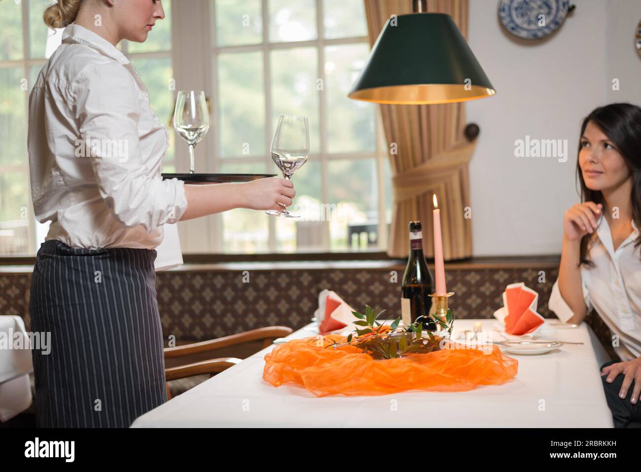 Weibliche Kellnerin, die einer attraktiven jungen Frau, die an einem formellen Tisch sitzt, Wein in eleganten Gläsern serviert Stockfoto